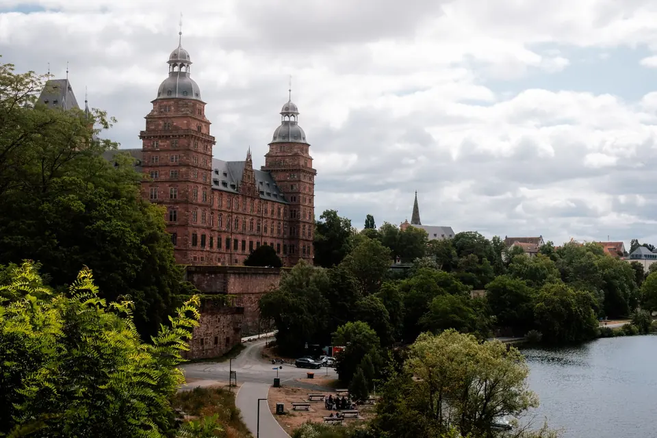 Malerische Ansicht von Schloss Johannisburg und dem Fluss in Aschaffenburg, Deutschland, umgeben von üppigem Grün.